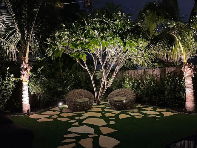 Lit backyard with two chairs, trees, and stone path at night.