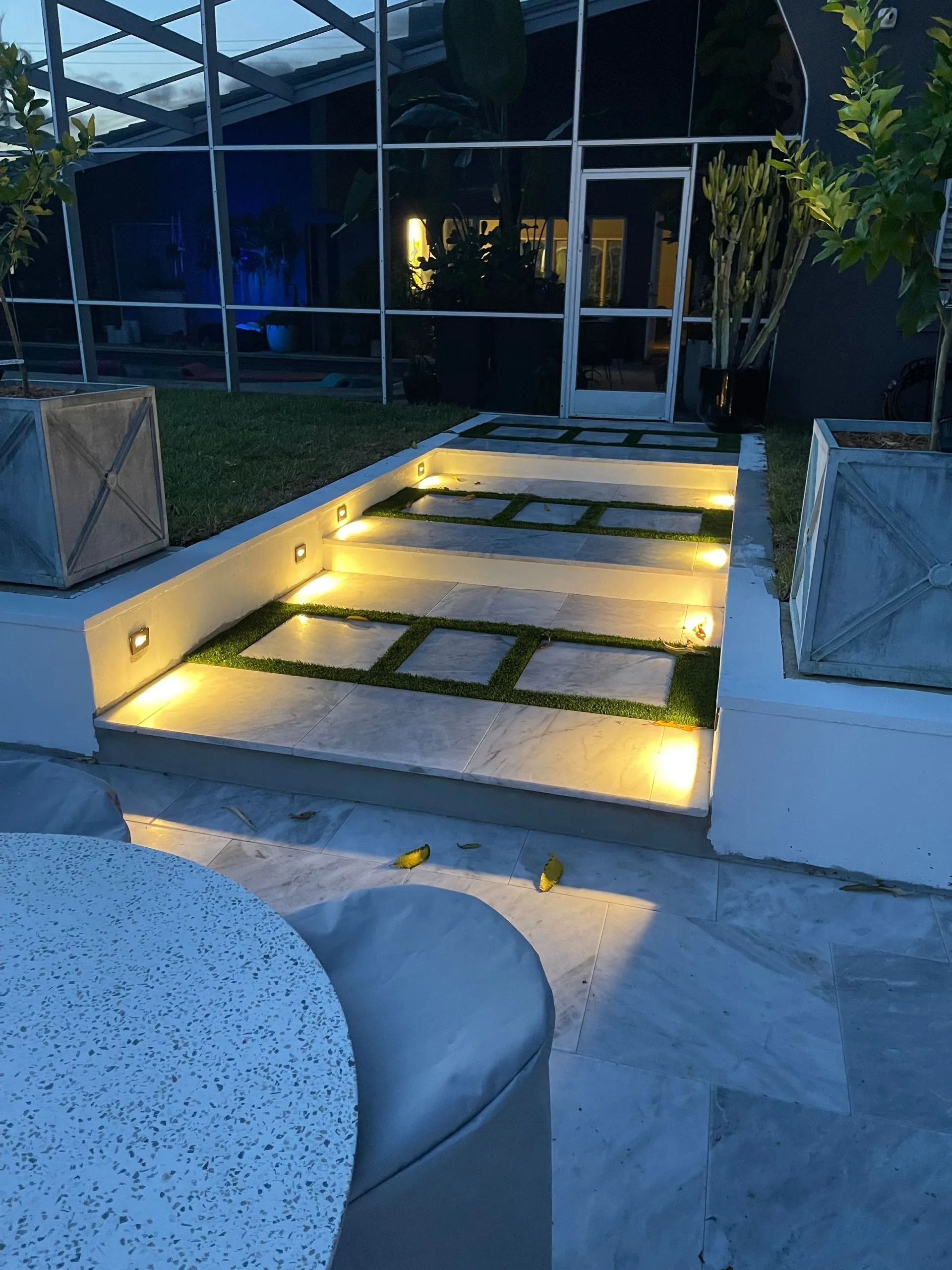 Outdoor staircase with built-in lighting and grass accents leading to a glass-enclosed area, at dusk.