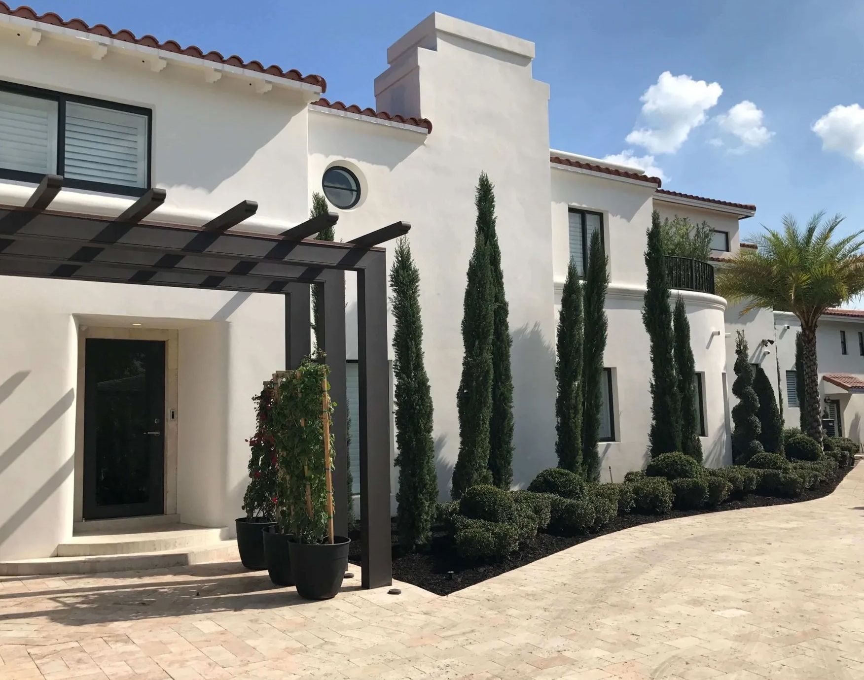 White stucco building with pergola, tall trees, and manicured landscaping.