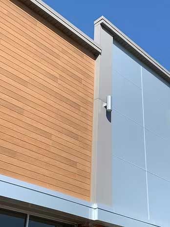 Building exterior with light brown wood paneling and silver metal siding, under a clear blue sky.