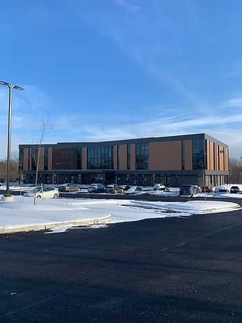 Modern two-story building with large windows and brown paneling, in a snowy parking lot under a blue sky.