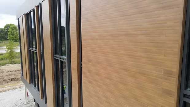 Exterior view of a building with wooden panels and large windows. The building is light brown with black window frames.