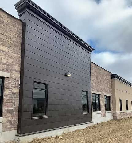 Dark metal paneling on a building exterior with small windows, brick and beige walls, and a light fixture.