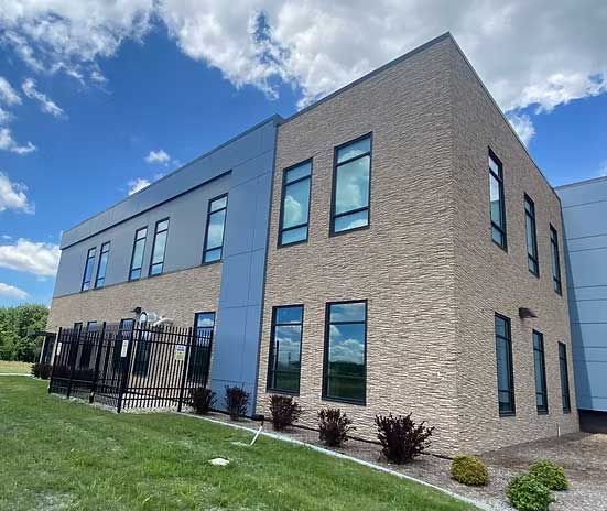 Two-story building with textured tan brick and blue metal siding. Dark-framed windows reflect the sky. Black fence and green grass.