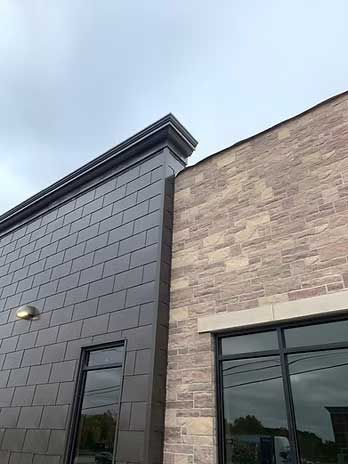 Exterior view of two-tone building. Dark grey, tiled facade adjoins a tan brick wall.