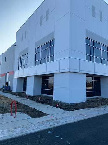 Corner of a modern, light-colored industrial building with large windows and a paved area.