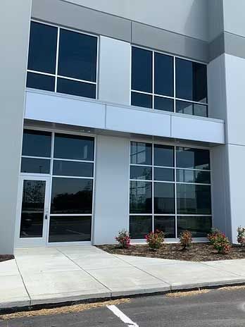 Exterior of a modern building with large windows and a glass door; concrete walkway and small flower bushes.