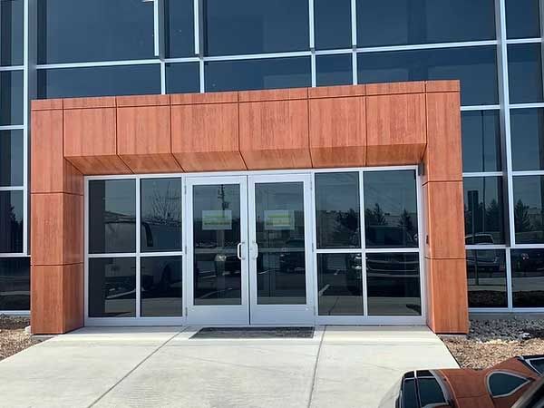 Building entrance with wood-toned frame, glass doors, and windows; a concrete walkway leads up to the door.