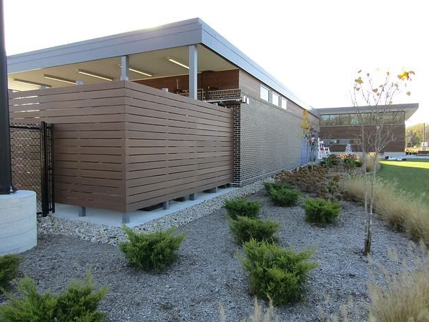 Modern building exterior with brown horizontal slat fence, green bushes, and gravel landscaping.