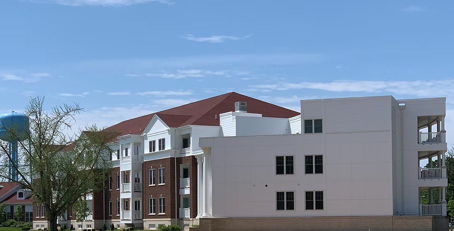 Multi-story building with a red roof, white walls, and a water tower in the background under a blue sky.
