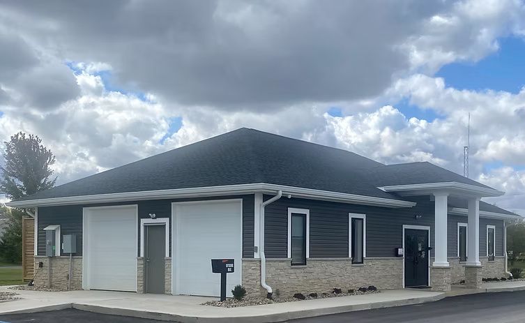 Exterior view of a one-story building with a dark gray roof and siding, two garage doors, and a black door.