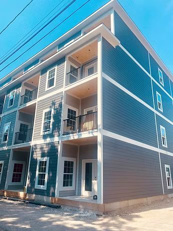 Three-story apartment building with gray siding, white trim, balconies, and clear blue sky.