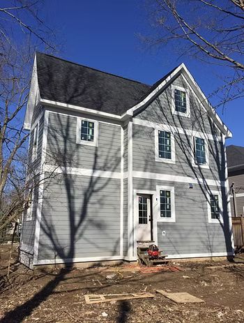 Two-story gray house under construction with white trim, windows, and black roof on a sunny day.
