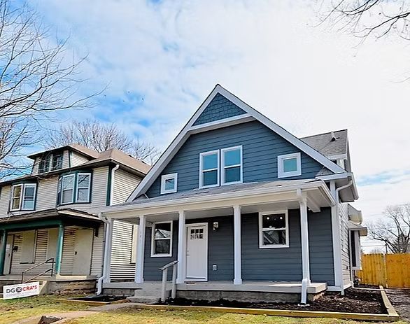 Blue house with white trim and porch, next to a beige house, cloudy sky.
