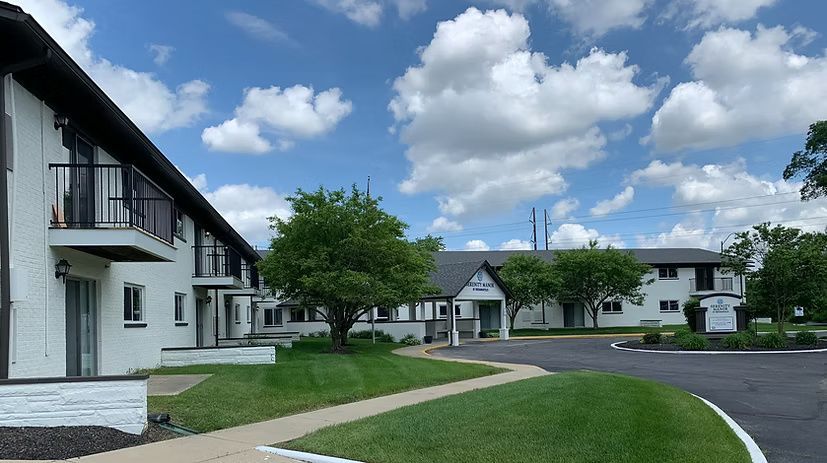 White apartment buildings with black trim under a blue sky with fluffy clouds; green grass and a paved driveway.