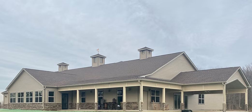 Beige house with a long porch, brown roof, and chimneys against a cloudy sky.