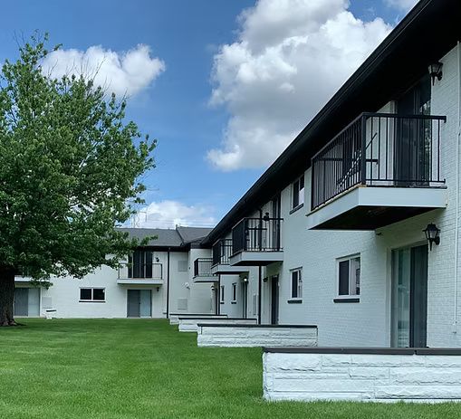 White brick apartment building with black trim and balconies, green lawn, blue sky.