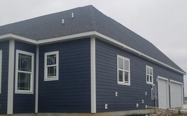 Blue house with white trim, windows, and garage doors under a dark gray roof. Cloudy sky.