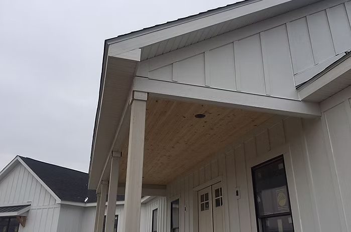 White farmhouse exterior with a covered porch and wood-paneled ceiling, under overcast skies.