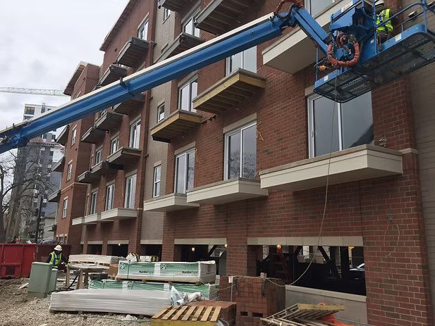 Construction site with a cherry picker extending towards a brick building with balconies. A worker stands below.