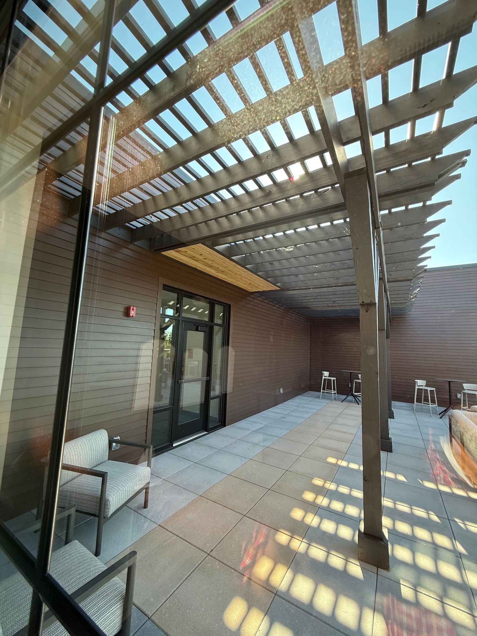 Covered patio with pergola slats, tiled floor, glass doors, and a narrow strip of sunlight against a brick wall