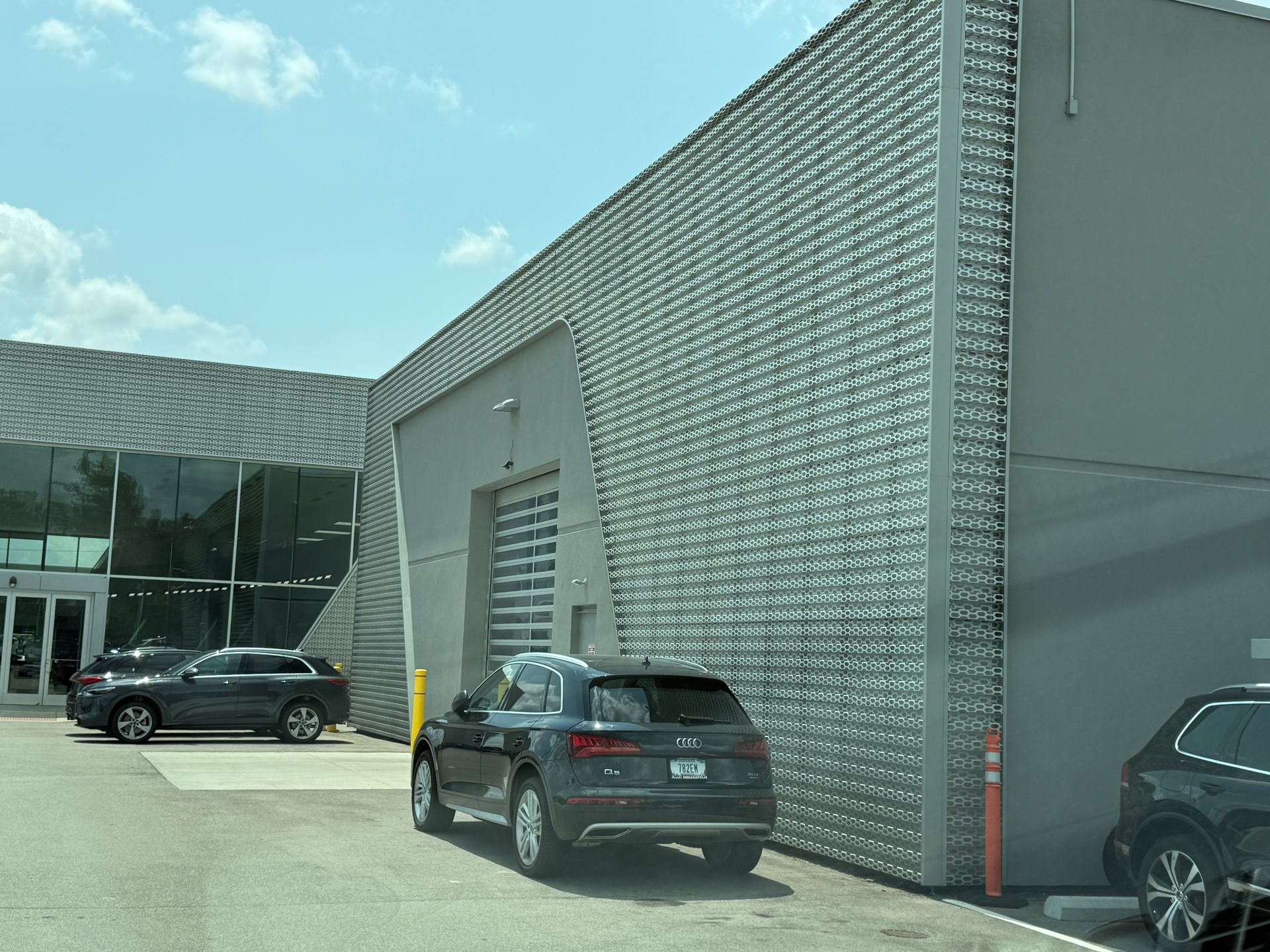 Parking lot outside a modern brick building, with several cars and a glass entrance under a blue sky.
