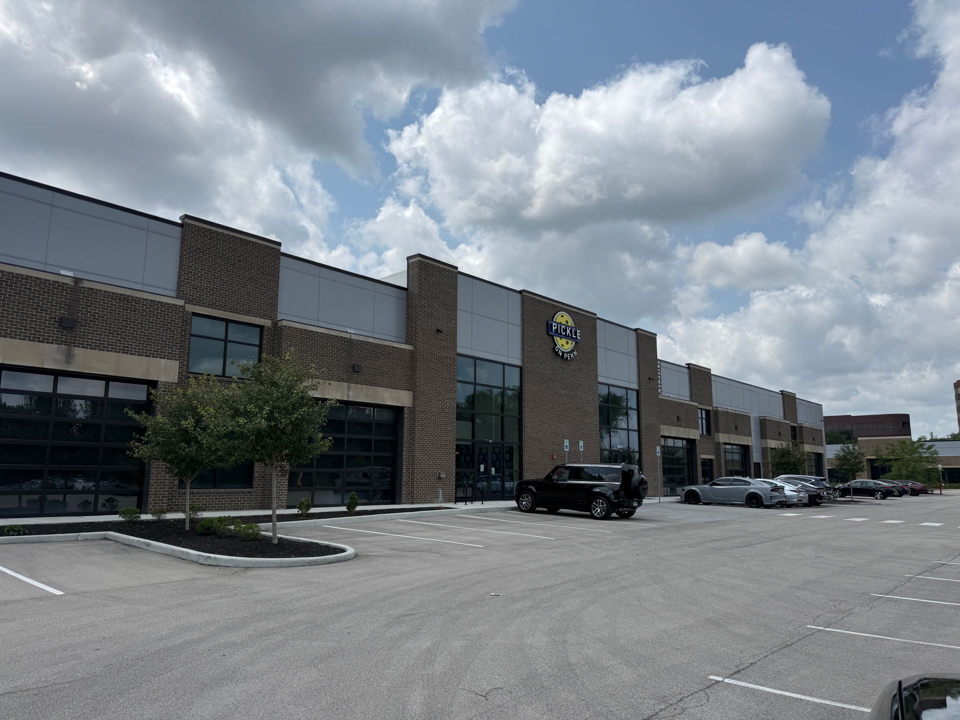 Strip mall storefronts with parked cars under a cloudy sky