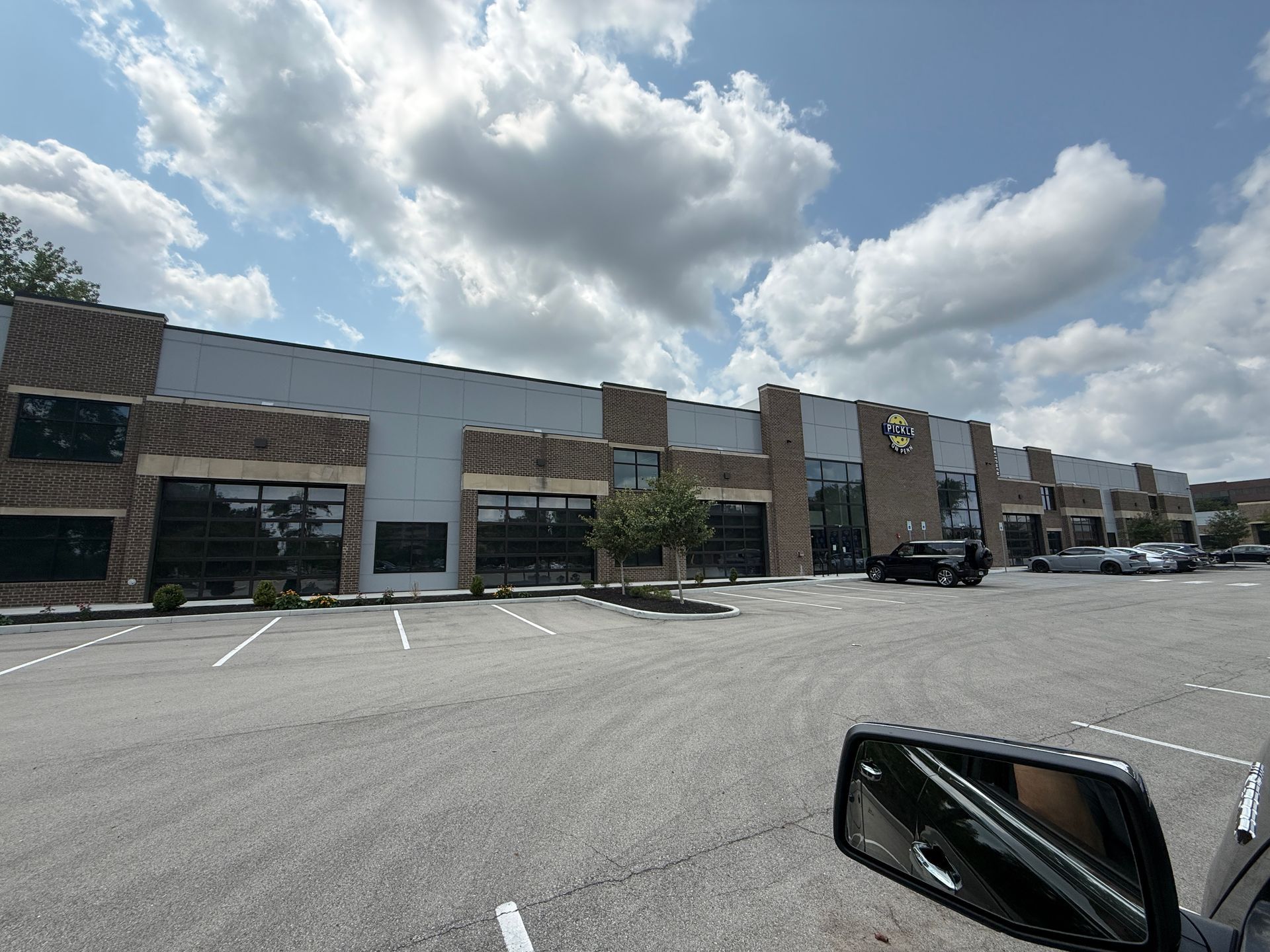 Shopping center storefronts and parking lot under a cloudy blue sky, seen from a car.