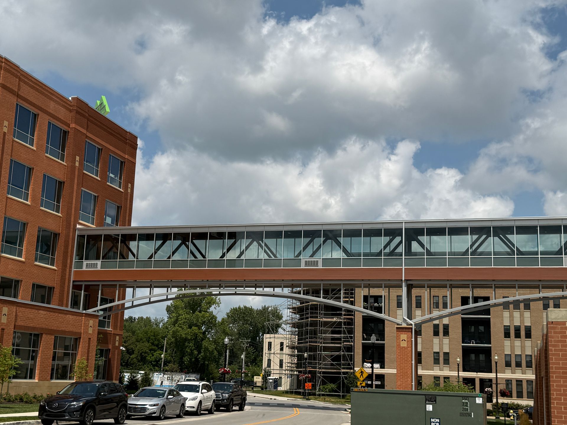 Brick campus buildings linked by a glass skybridge under cloudy skies, with cars parked below.