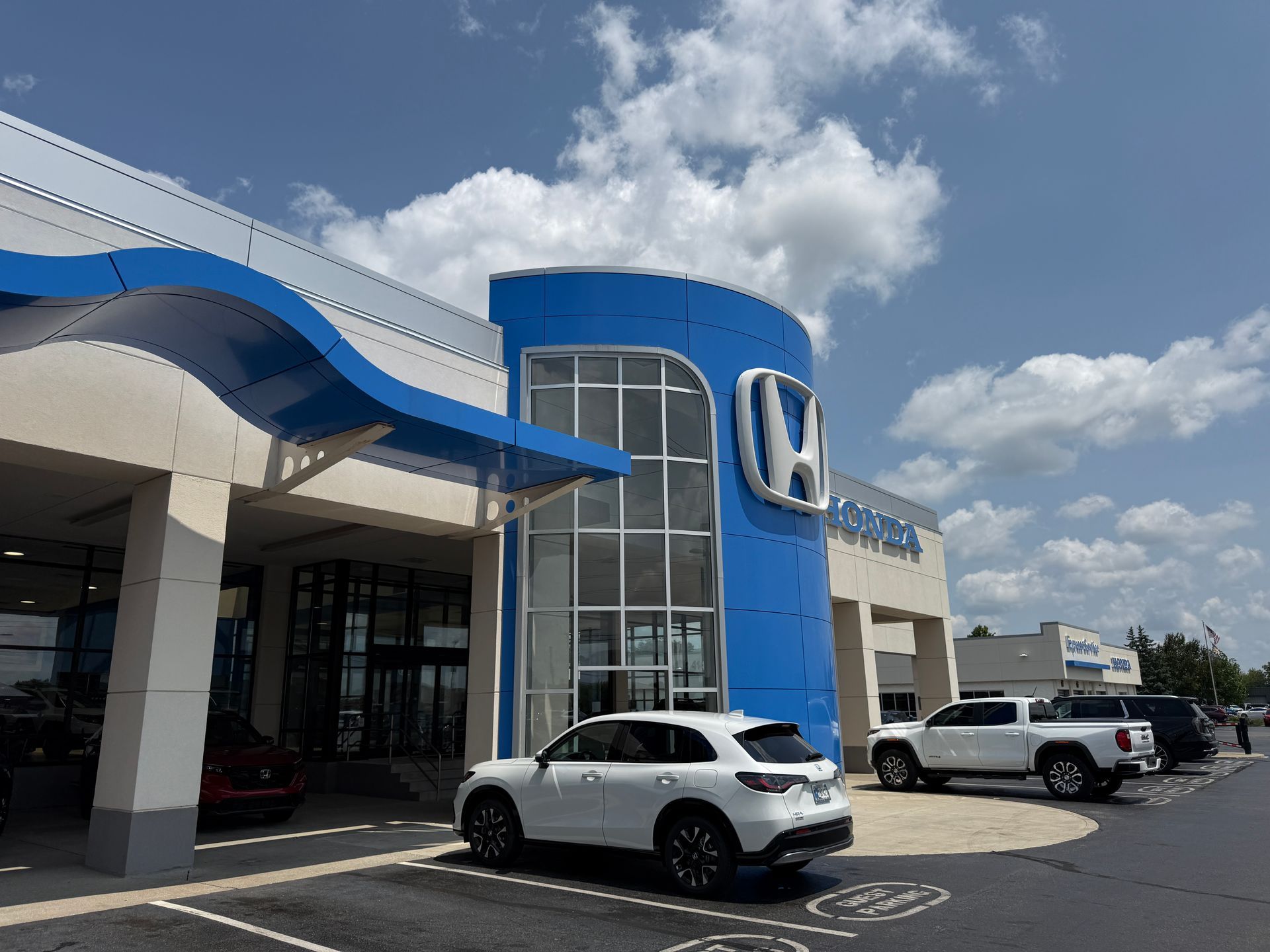 Exterior of a Honda dealership with blue accents, glass entrance, and parked cars under a partly cloudy sky