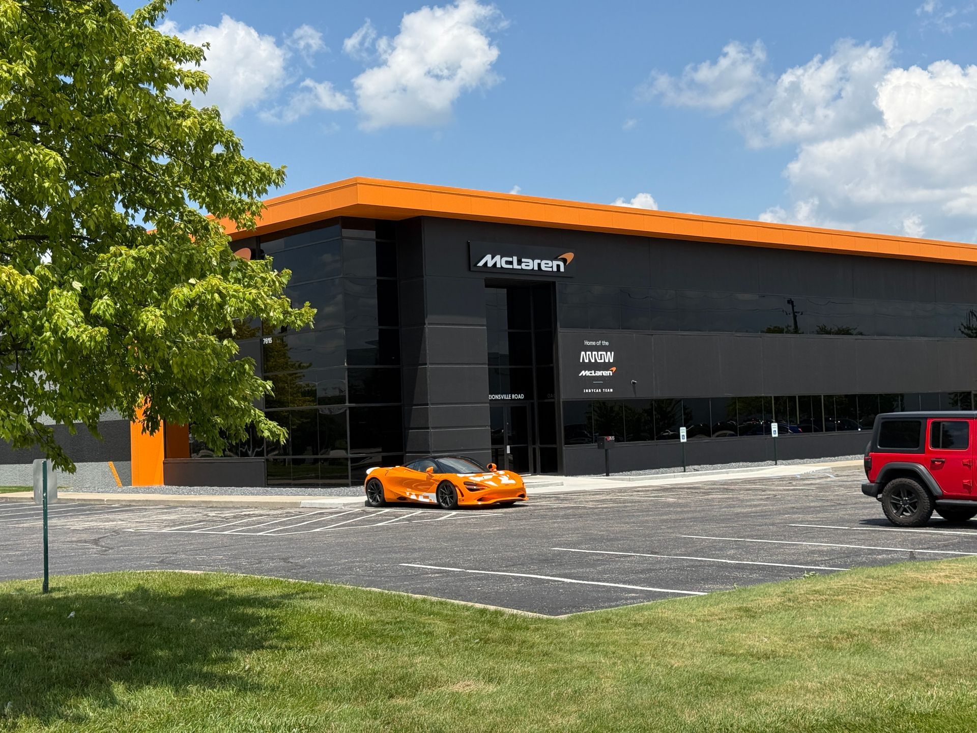 Orange sports car parked outside a black building with orange trim, beside a red SUV and trees.