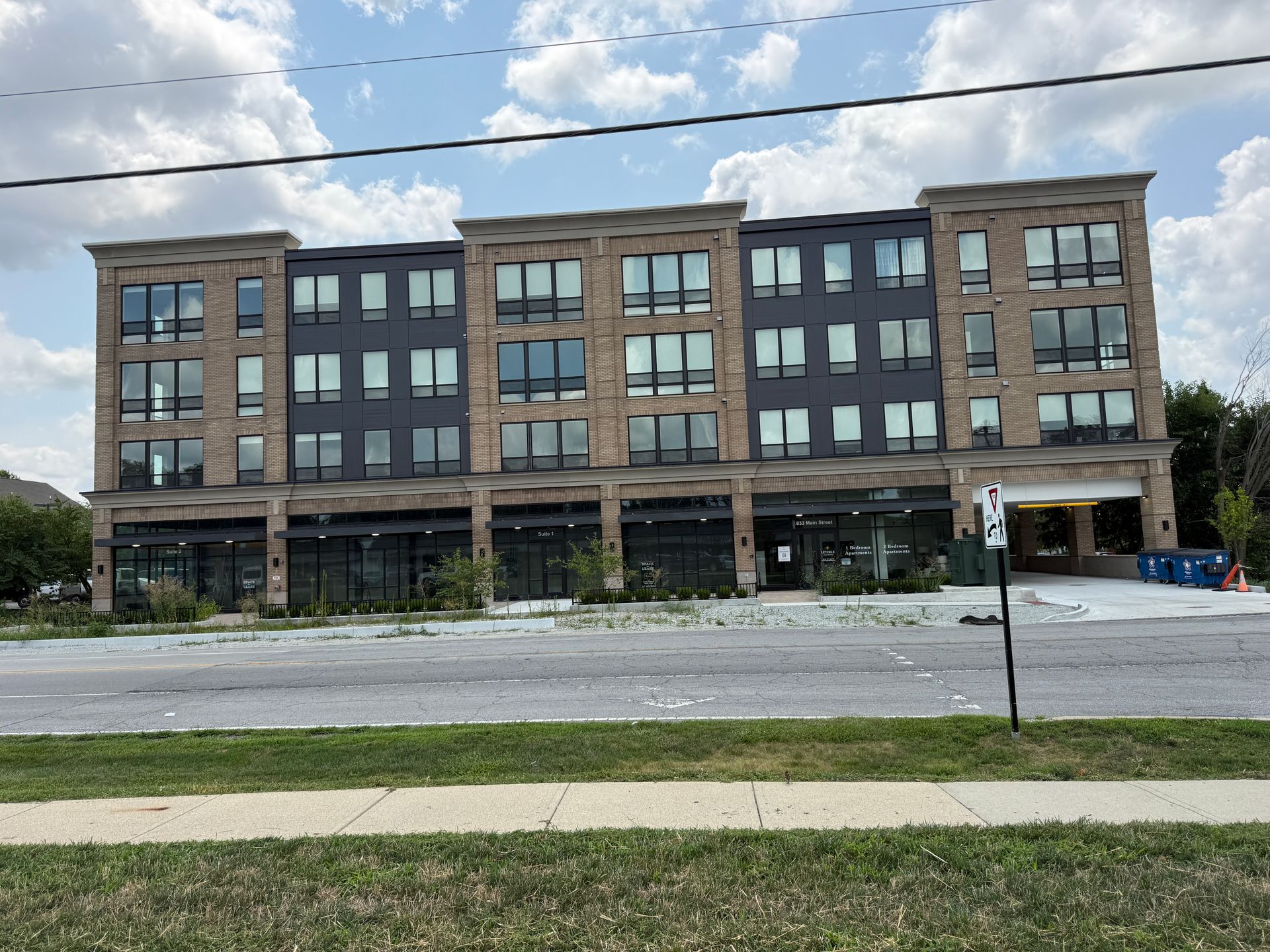 Modern four-story brick apartment building with large windows and a grassy foreground under a cloudy sky