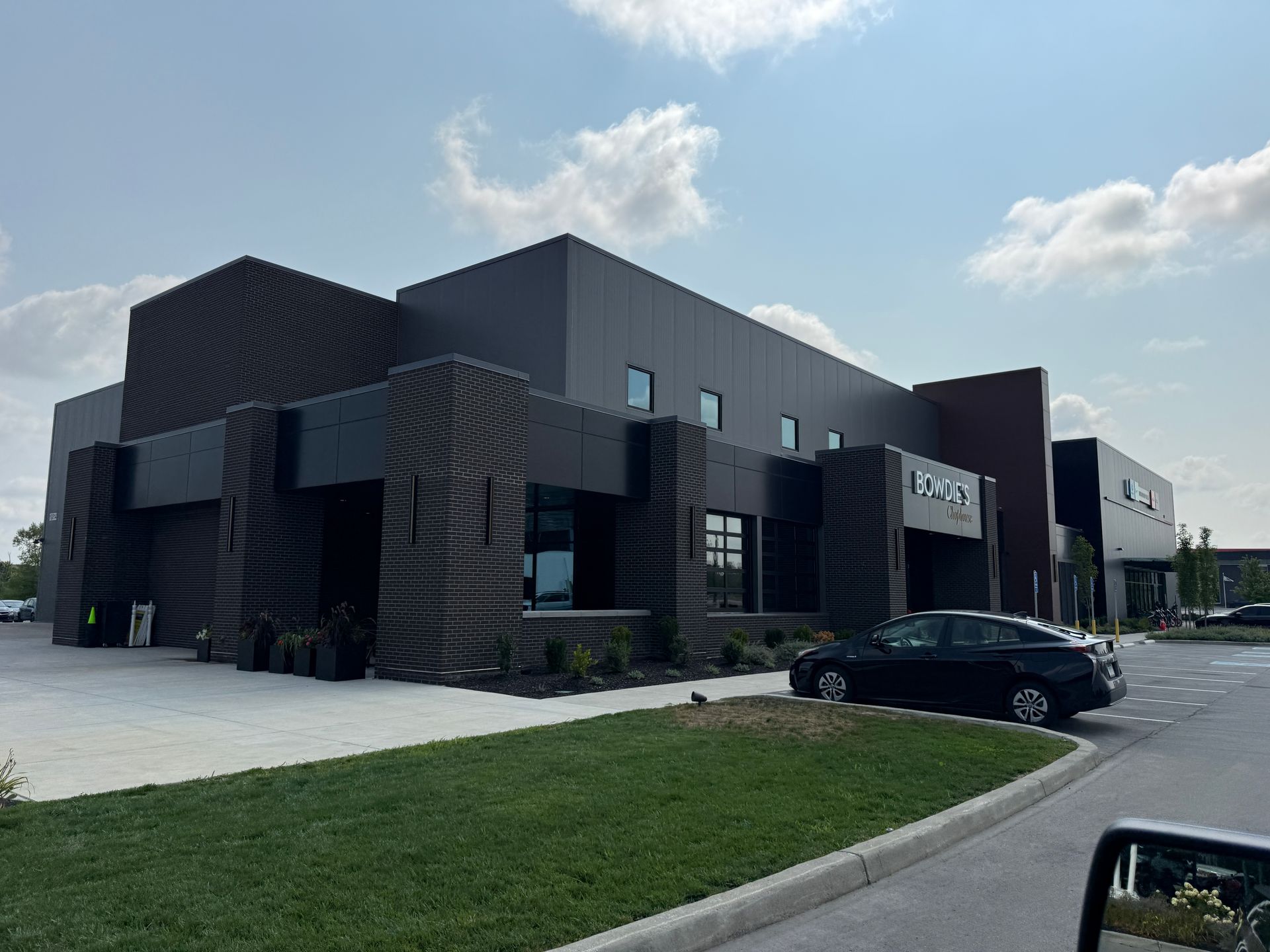 Modern black commercial building with a parked car and grassy curb under a partly cloudy sky