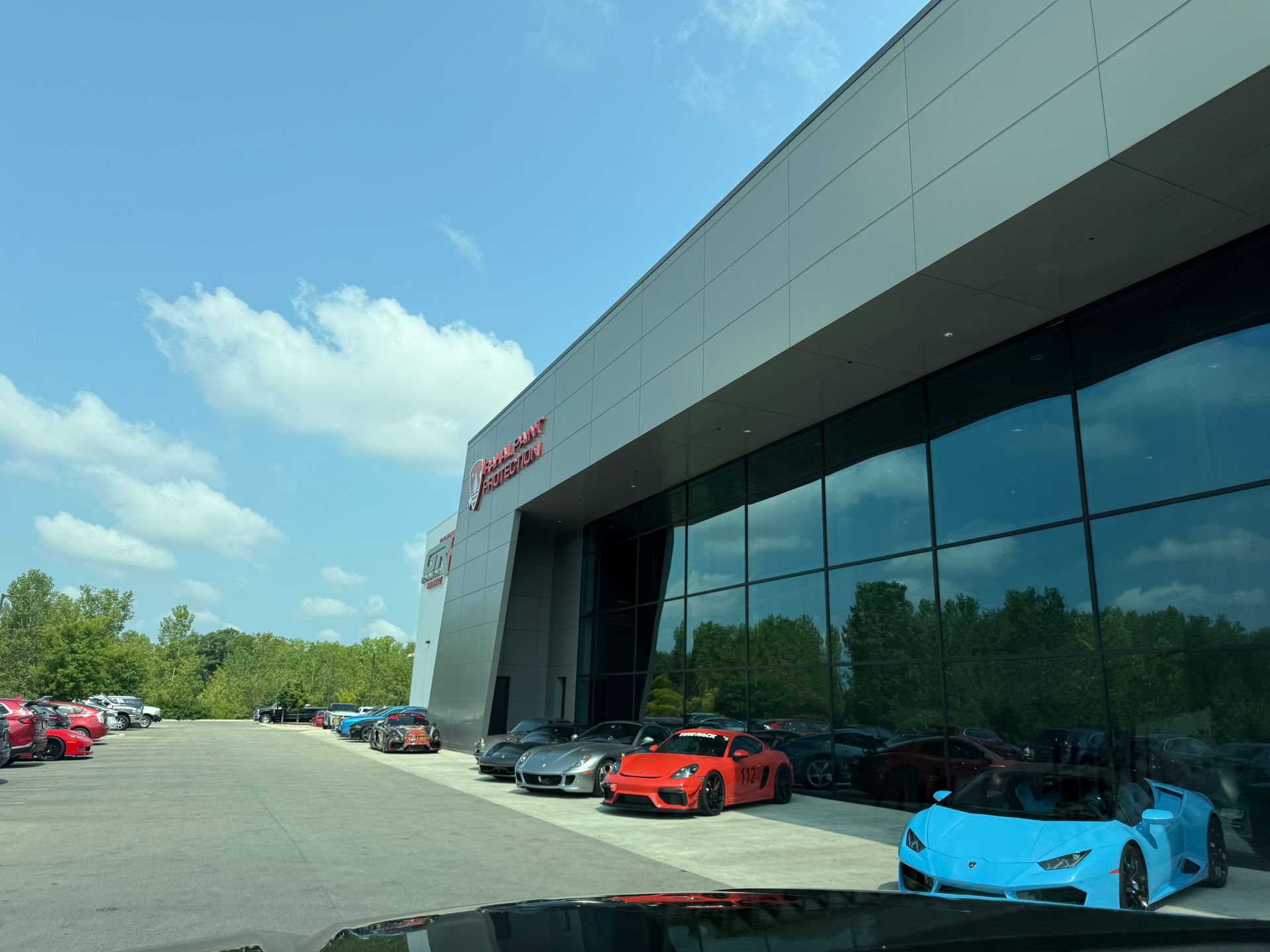 Sports cars lined up outside a modern dealership with reflective windows and a partly cloudy sky.