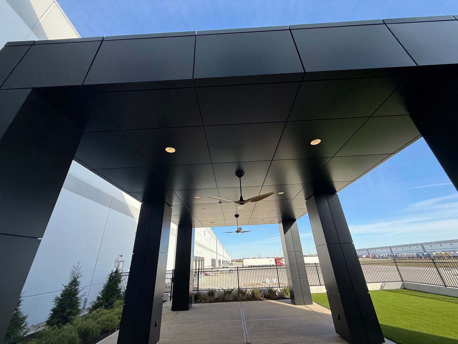 Modern covered walkway with black columns, ceiling lights, and a view of a fence and blue sky