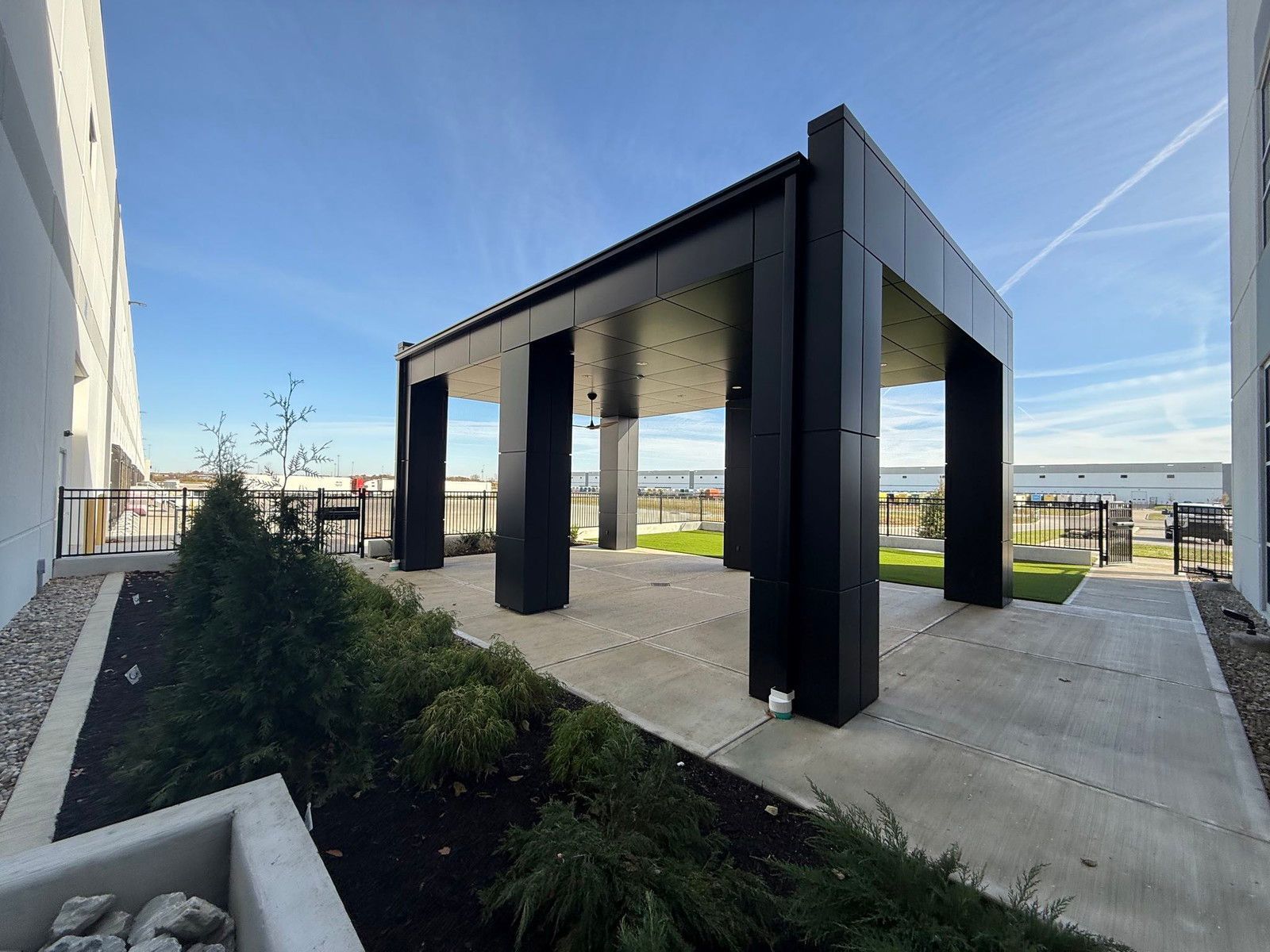Modern black pavilion with square columns on a concrete patio beside a building, under a blue sky