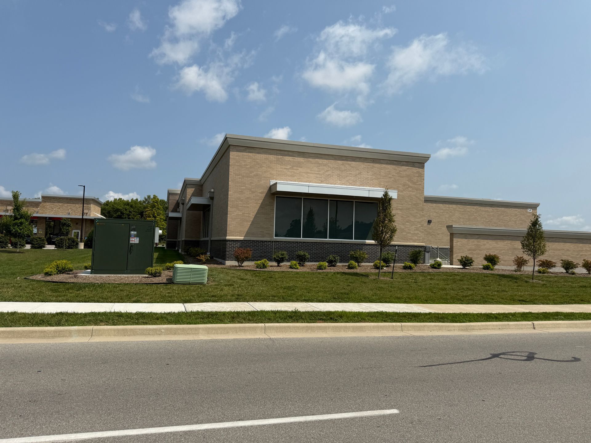 Modern beige building with large windows, landscaped front lawn, and road in the foreground under a blue sky
