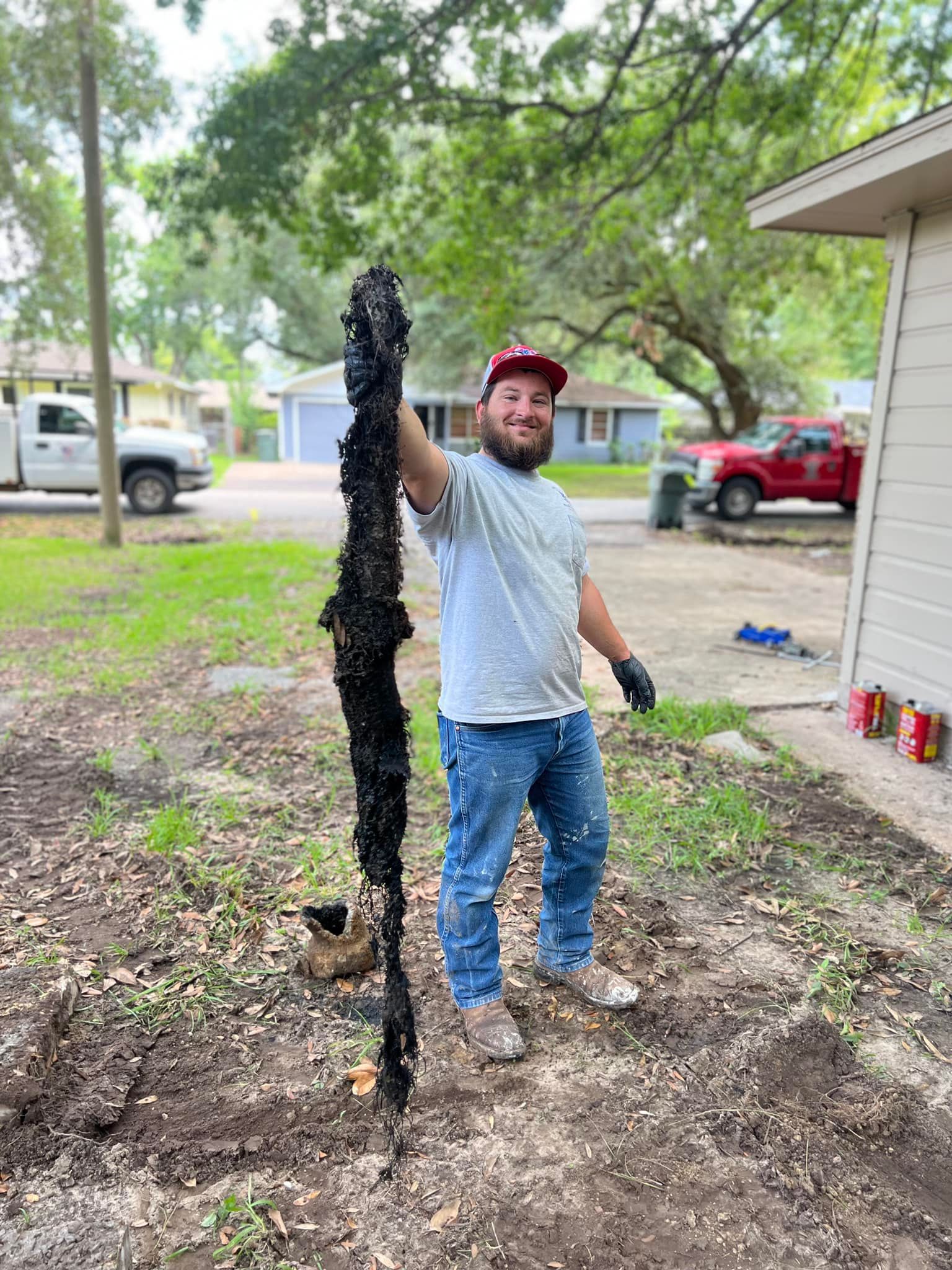 Man holding long, dark, debris item in a yard. Smiling, wearing a hat, jeans, and gloves. House and truck in background.