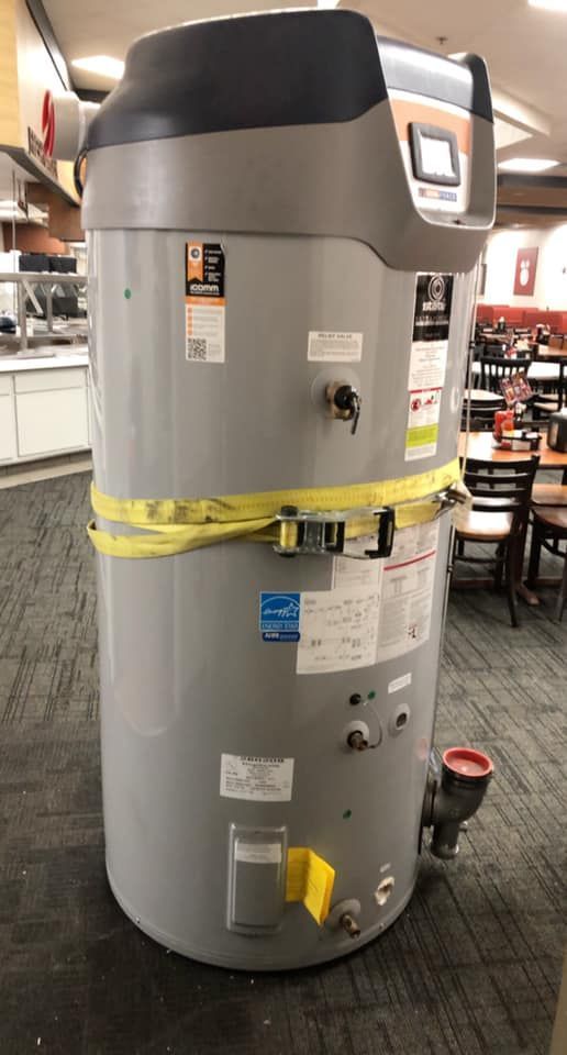 Tall gray water heater with black top, yellow strap, and red pipe in a public area.