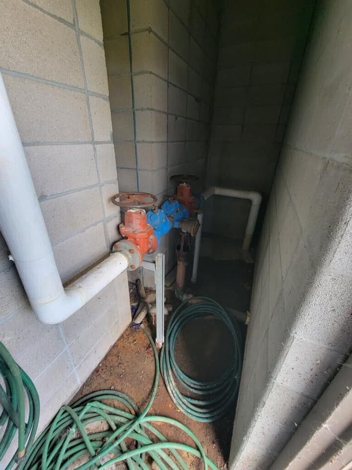 Water valves and pipes in a small concrete enclosure with a coiled green hose on the floor.