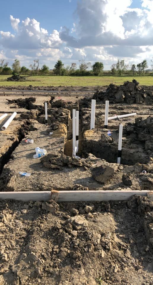 Construction site with PVC pipes emerging from dirt. Blue sky, clouds, and a grassy field in the background.