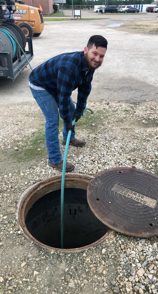 Man using a hose to work in an open manhole. He's smiling. Outdoors with equipment visible.