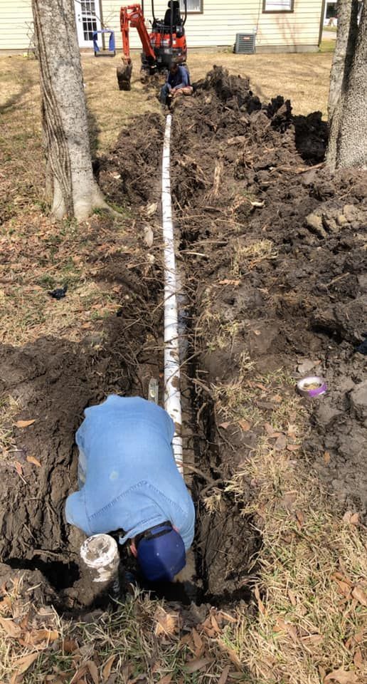 Man in blue shirt working on white pipe in a trench, orange tractor in the background.