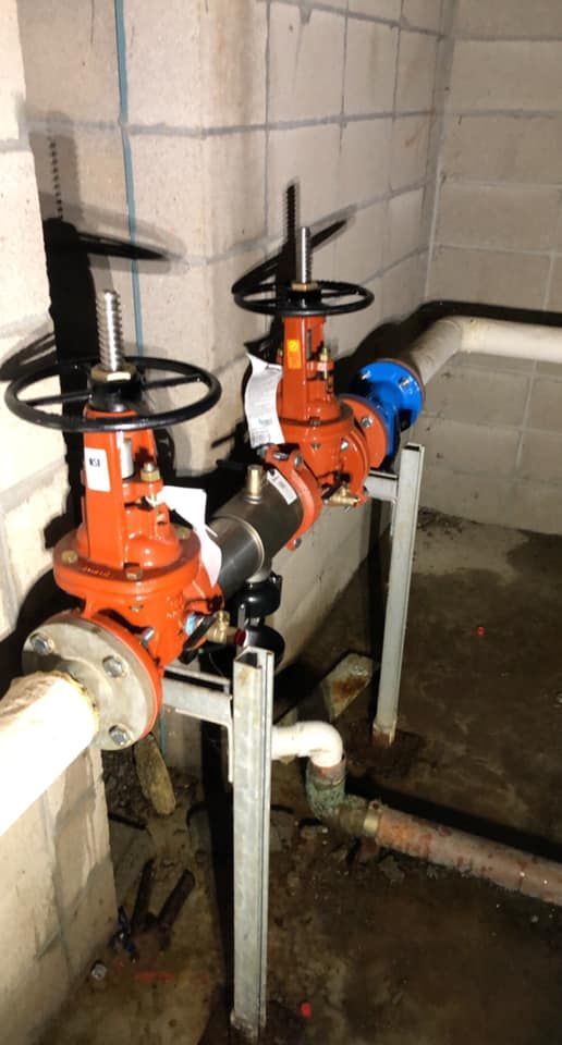 Two red water valves with black handles and connected piping in a concrete-walled basement.