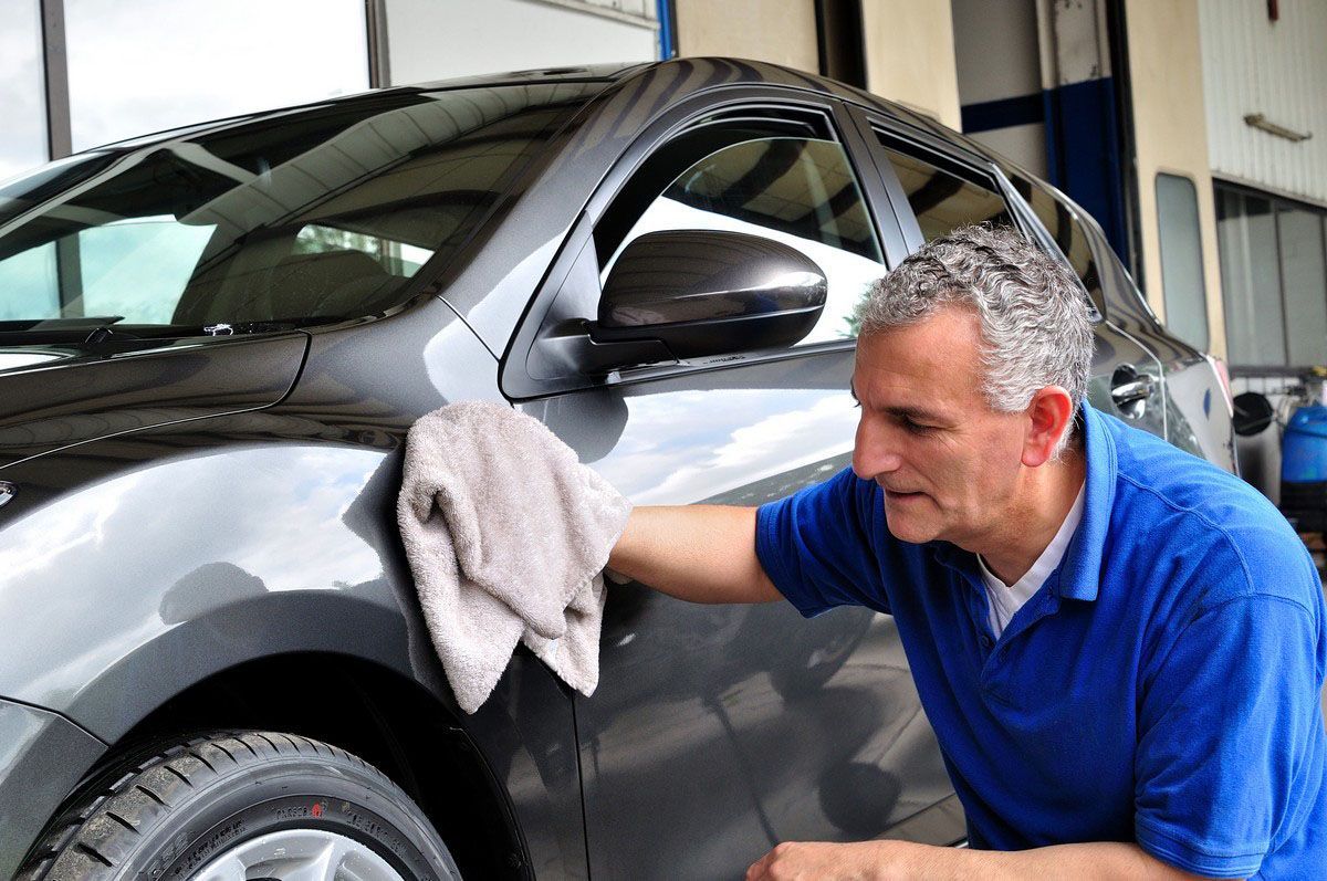Man in blue shirt wiping a dark gray car with a tan cloth in a garage.
