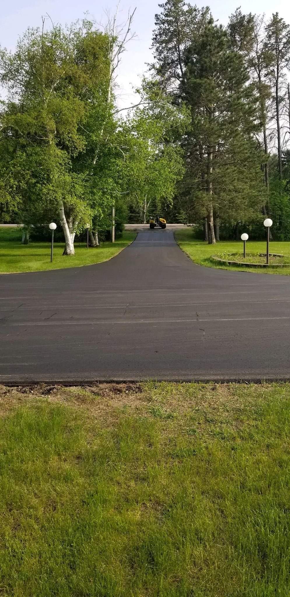 A black asphalt driveway leading to a house surrounded by trees and grass.