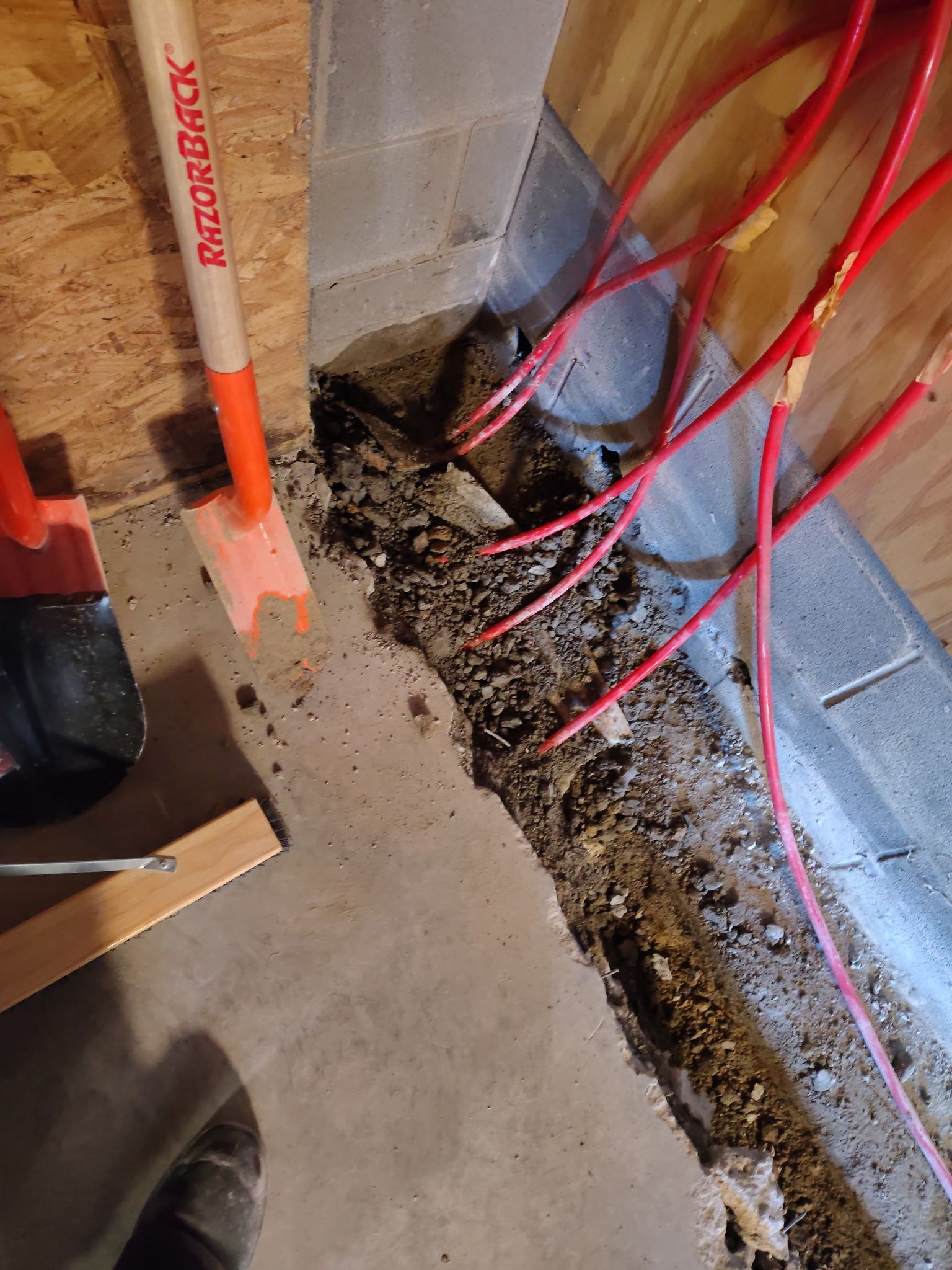 Trench in concrete floor with red pipes.  Pipes enter wooden wall.  Tool on wood floor next to trench.