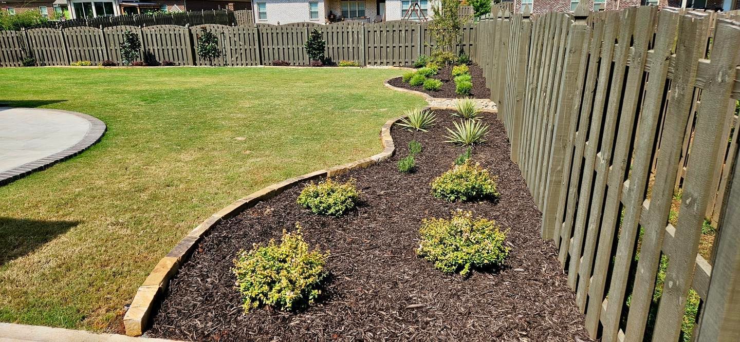 A backyard with a grassy lawn, a flower bed with mulch and bushes, and a wooden fence.