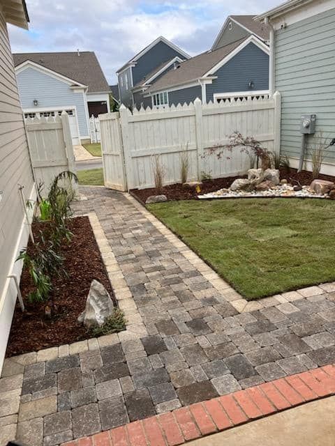 Brick path leads to a backyard with a small lawn, white fence, and houses in the background.