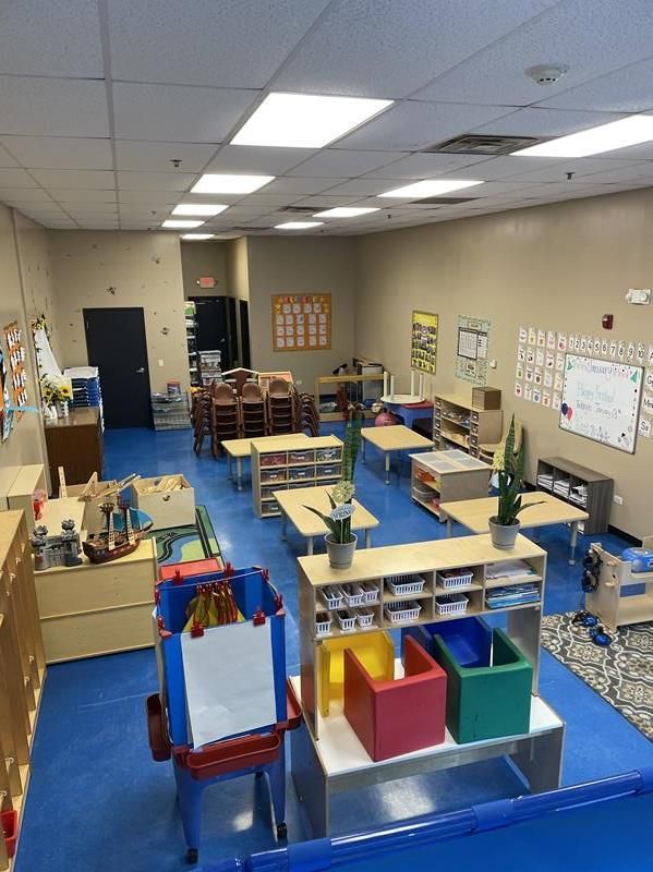 A brightly lit preschool classroom with blue floors, small tables, colorful chairs, learning materials, and wall decorations.